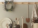 En minimalistisk køkkenvæg med køkkenredskaber af træ, en ternet tallerken, en potteplante, en hængende krans, et håndklæde og dekorative genstande, der er pænt arrangeret på en beige baggrund.
