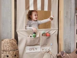 Et smilende barn står bag et legestativ af stof med stribede detaljer og holder en legetøjscupcake. Stativet har lommer mærket OPEN fyldt med plyslegetøj.