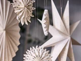 Papirdekorationer i forskellige geometriske former, bl.a. stjerner og rosetter, hænger i snore mod en sløret baggrund. Dekorationerne er hvide med fine udskæringsmønstre.