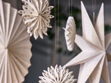 Papirdekorationer, bl.a. stjerner og snefnug, hænger i snore. Dekorationerne er hvide og kunstfærdigt foldede, hvilket skaber en festlig og delikat atmosfære.