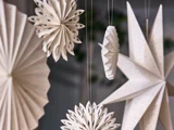 Papirdekorationer, bl.a. stjerner og snefnug, hænger i snore. Dekorationerne er hvide og kunstfærdigt foldede, hvilket skaber en festlig og delikat atmosfære.