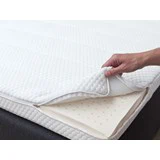 En hånd løfter det øverste lag af et hvidt madrasbetræk og afslører et lag af perforeret skum nedenunder.