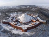 Luftfoto af en stor cirkulær fæstning med røde murstensvægge og et kuppeltag, omgivet af snedækkede træer og landskab om vinteren.