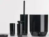 Et sæt skinnende sort badeværelsestilbehør, herunder en toiletbørsteholder, en sæbedispenser, en tumbler og en affaldsspand, arrangeret på en lys overflade mod en almindelig baggrund.