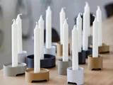 Flere hvide bloklys er placeret i moderne, runde lysestager i forskellige neutrale farver, herunder grå, sort og guld, arrangeret på en lys træoverflade.