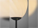 Nærbillede af en moderne gulvlampe med en hvid, struktureret, kugleformet lampeskærm og en sort fod mod en grå baggrund.