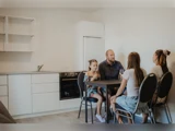 En familie på fire sidder omkring et lille bord i et moderne køkken og snakker og smiler. Rummet har hvide skabe og minimal indretning.