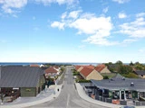 En lige vej fører mod havet, omkranset af huse med røde og gule tage under en delvis overskyet blå himmel.