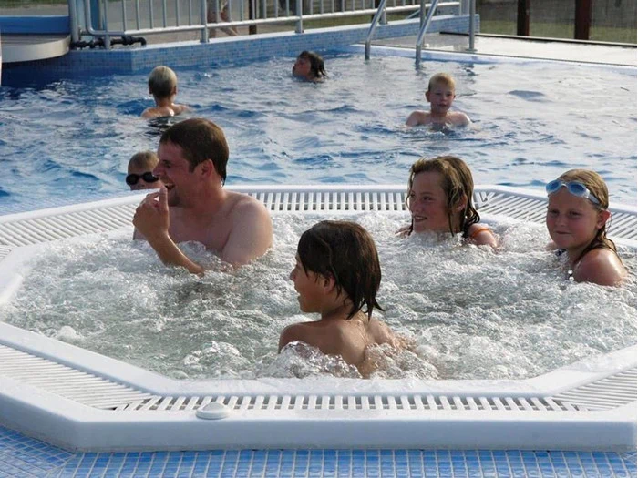 Flere børn og en voksen sidder i et boblende spabad, mens andre svømmer og leger i poolen omkring dem på en solrig dag.