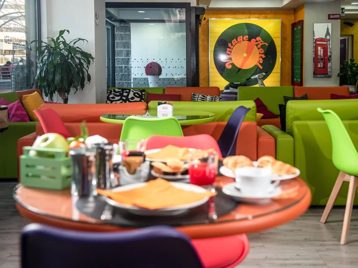 En farverig café med lyse stole og sofaer, et bord med morgenmadsretter og et stort Vintage Hotel-skilt på bagvæggen.