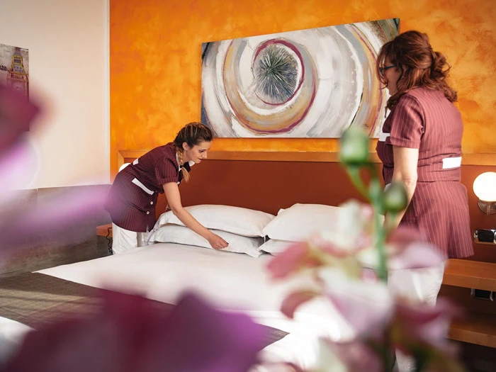 To stuepiger i uniformer reder en pænt opredt seng i et lyst indrettet hotelværelse med orange vægge og abstrakt kunst over hovedgærdet.