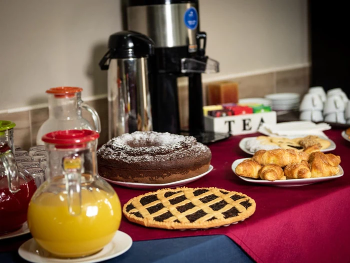 En morgenmadsbuffet med juicekander, en kaffeurne, en chokoladekage, en frugttærte, croissanter og teposer på et bord med en rød dug.