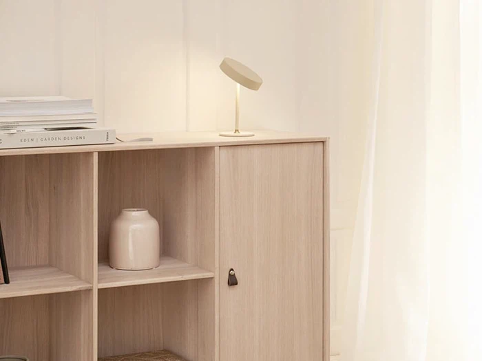 Et minimalistisk træskab med hylder, en beige bordlampe, stablede bøger og en lys keramikvase mod en hvid væg med gennemsigtige gardiner.