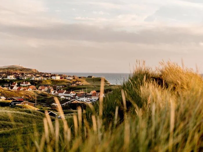 En kystby med huse med røde tage, grønne græsklædte bakker i forgrunden og havet synligt under en overskyet himmel i baggrunden.