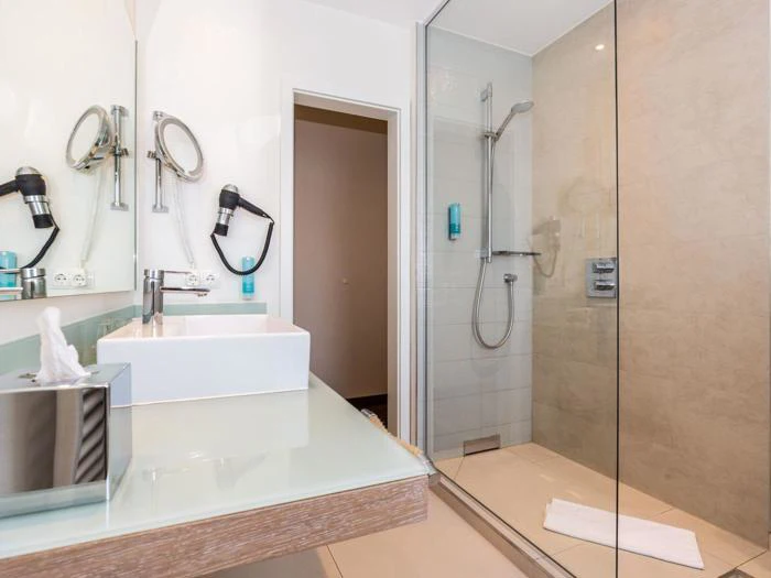 Moderne badeværelse med glasbruser, væghængt brusehoved, hvid håndvask, spejl, hårtørrer, papirkurv og et håndklæde på gulvet uden for bruseren.