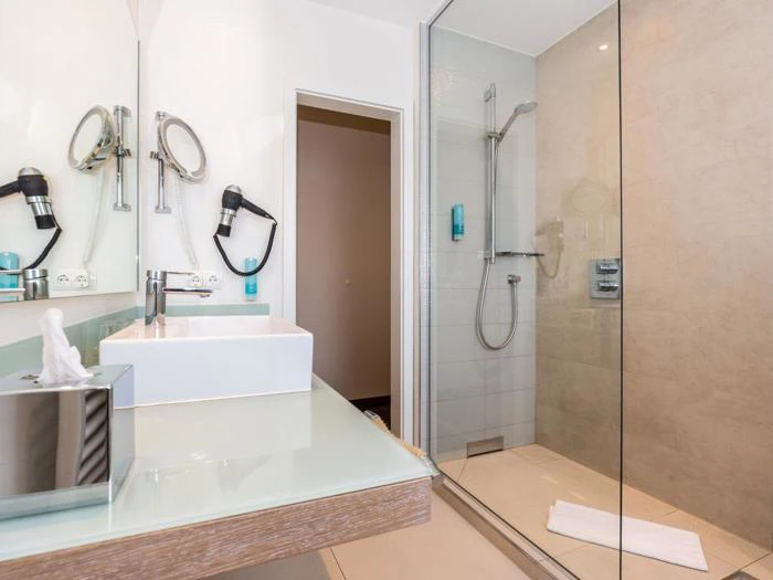 Moderne badeværelse med glasafskærmet bruser, væghængt brusehoved, vaskeskab med håndvask, papirkurv og væghængt hårtørrer. Der ligger et hvidt håndklæde på gulvet.