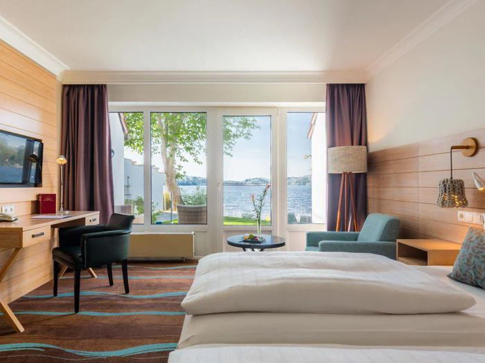 Lyst hotelværelse med to senge, skrivebord, stol, lænestol og store vinduer med udsigt til træer, gårdhave og en sø udenfor.