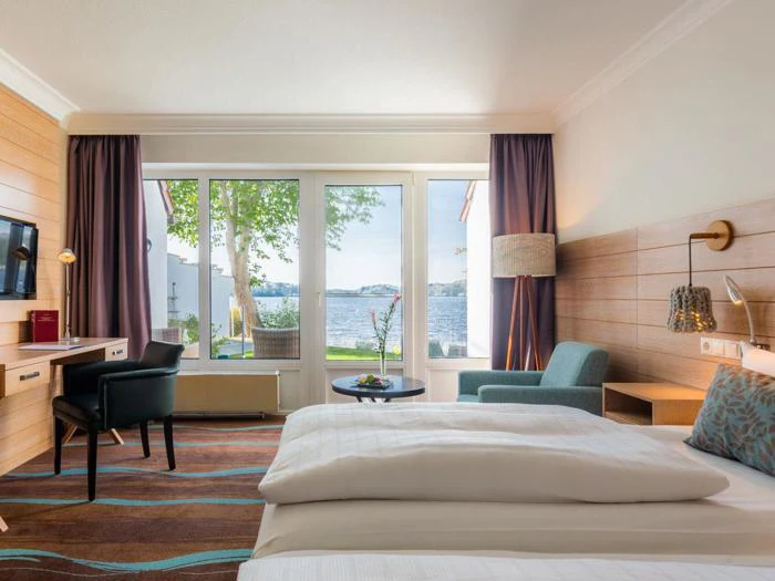 Moderne hotelværelse med to senge, et skrivebord, en lænestol og store vinduer med udsigt til en sø og træer udenfor.