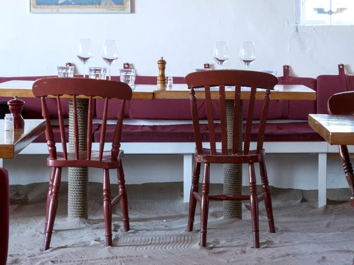 To tomme træstole ved et bord med vinglas og vandglas, inde i en restaurant med sand på gulvet og rødbrune stole.