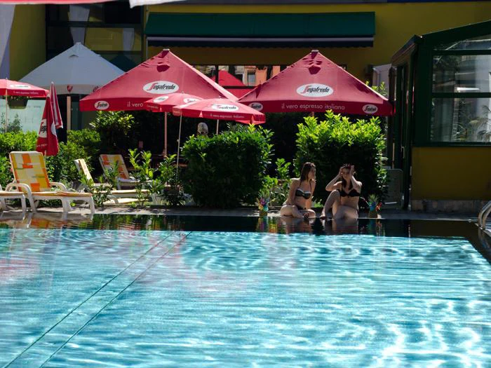 To kvinder i bikini sidder og taler ved en pool med røde parasoller og liggestole i baggrunden på en solrig dag.