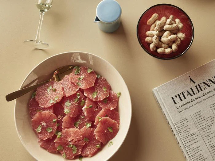 En tallerken med grapefrugt i tynde skiver med krydderurter, en skål med peanuts i skaller, et glas hvidvin, en lille blå kande og en del af en avis på et beige bord.
