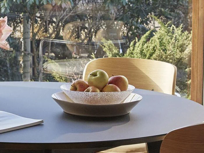 En klar skål fyldt med æbler og ferskner står oven på en hvid tallerken på et rundt, gråt bord med træstole og grønne planter i baggrunden.