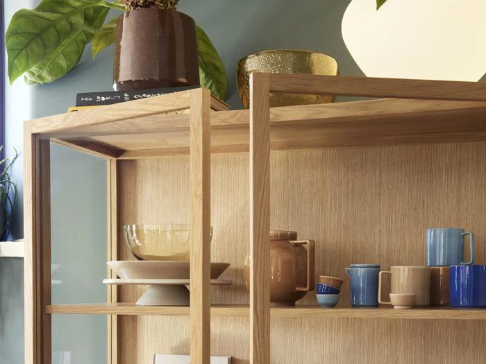 En træhylde med keramikskåle, krus, kopper og en tekande i neutrale og blå toner, med en potteplante og pyntegenstande på den øverste hylde.