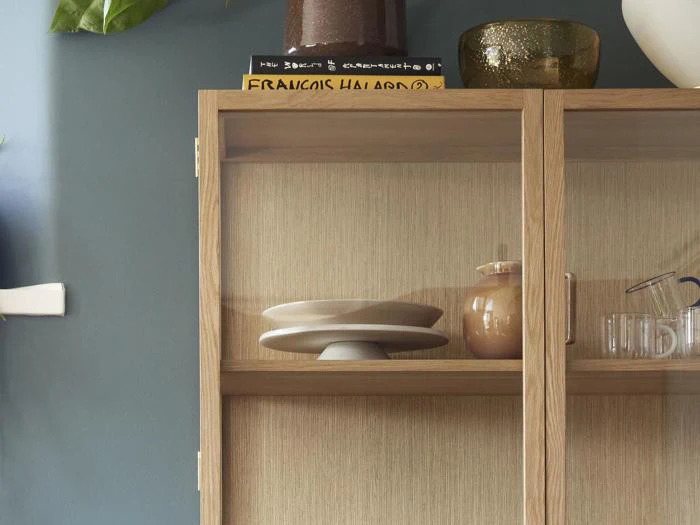 Et træskab med glasdøre viser tallerkener, en keramikkrukke og glasvarer. Dekorative genstande står øverst mod en blå væg med en bladplante delvist synlig til venstre.