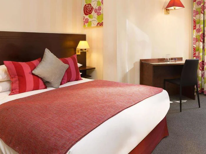 Et hotelværelse med en pænt redt dobbeltseng med røde og lyserøde puder, et rødt tæppe, et træskrivebord med en stol og blomstrede gardiner.