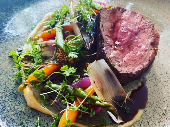 En tallerkenret med en tyk skive mediumstegt oksekød, garneret med mikrogrønt, asparges, gulerødder, rødløg og en cremet sauce, alt sammen kunstfærdigt anrettet på en struktureret tallerken.