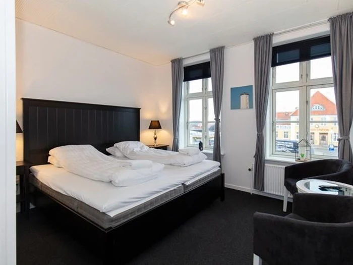 Et moderne hotelværelse med en dobbeltseng, to lamper, to vinduer med grå gardiner og to sorte stole omkring et glasbord.