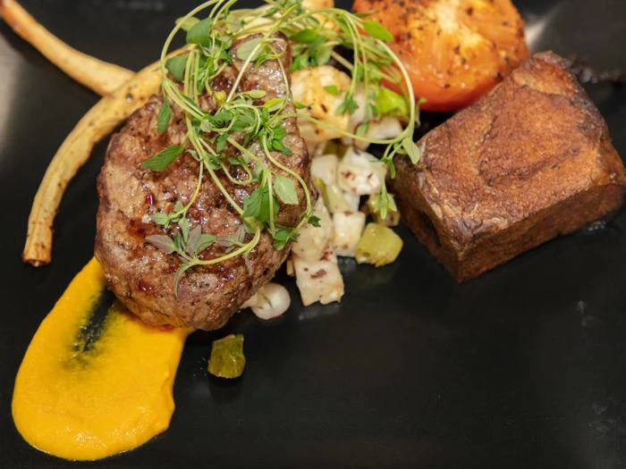 En gourmetret med en grillet bøf toppet med mikrogrønt, ristede grøntsager, en terning af sprød kartoffel og en klat lysegul sauce på en sort tallerken.