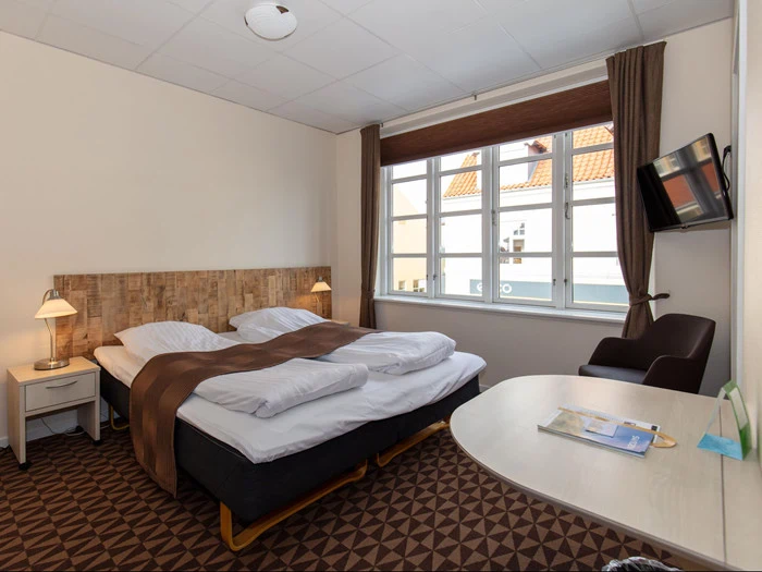 Et pænt hotelværelse med en dobbeltseng, brune tæpper, natborde med lamper, et stort vindue, et væghængt tv, en stol og et skrivebord.