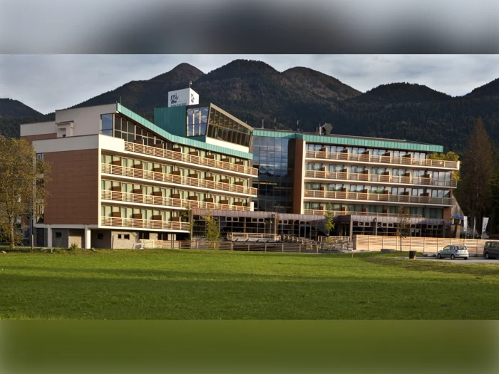 En moderne hotelbygning i flere etager med balkoner, der ligger på en baggrund af skovklædte bjerge og en græsmark i forgrunden.