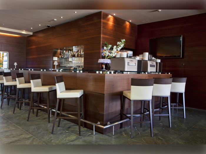 En moderne bar med trævægge og -disk, høje stole med hvide sæder, et stort tv, kaffemaskiner og et udvalg af flasker og glas på hylder bag baren.