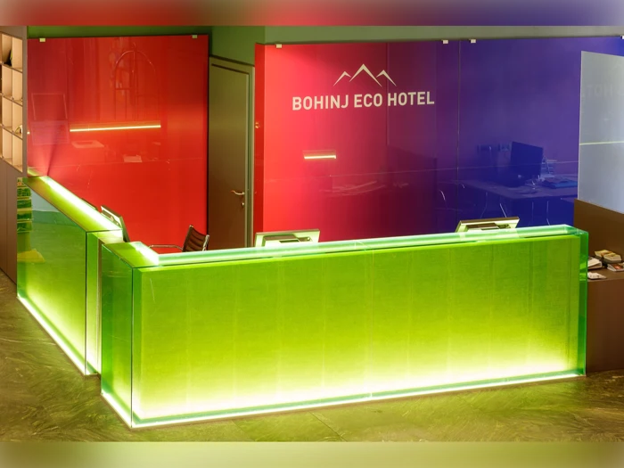 En moderne hotelreception med grøn belysning foran en rød væg med skiltet Bohinj Eco Hotel. To computerskærme står på disken.