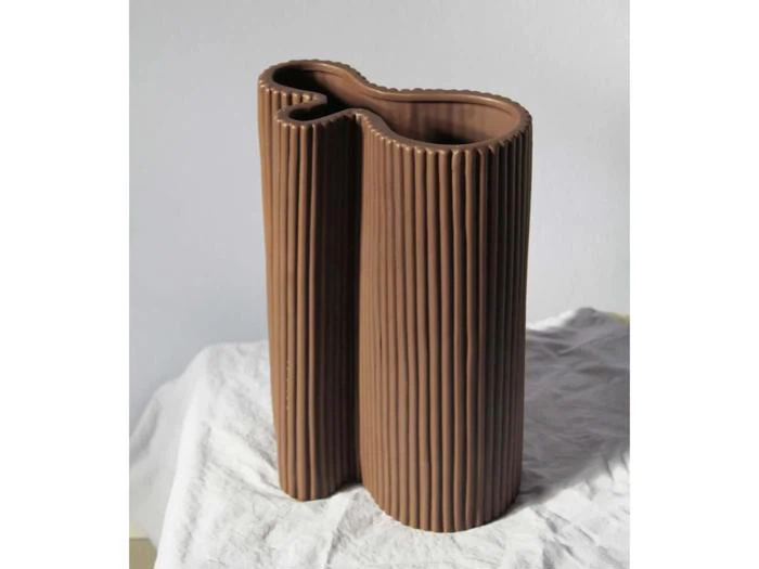 En ribbet, abstrakt brun keramikvase med en uregelmæssig bølget form står på et hvidt klæde mod en neutral baggrund.