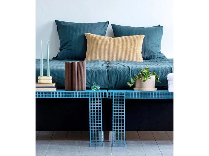 På en moderne bænk med blå gitterfliser står stearinlys, bøger, en grøn glaspynt, en potteplante og foldede håndklæder foran en seng med blågrønt sengetøj og en solbrun pude.