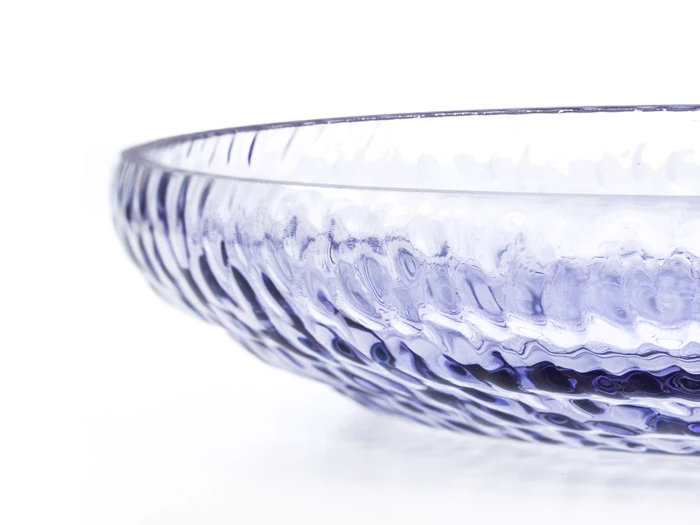 Et nærbillede af en gennemsigtig, struktureret glasskål med en diskret lilla nuance på en hvid baggrund.