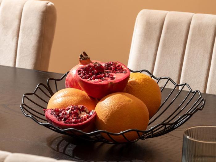 En frugtskål i sort tråd på et mørkt bord rummer to halve granatæbler og tre appelsiner med beige stole i baggrunden.