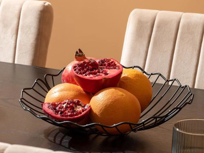 En frugtskål i sort tråd på et mørkt bord rummer to halve granatæbler og tre appelsiner med beige stole i baggrunden.