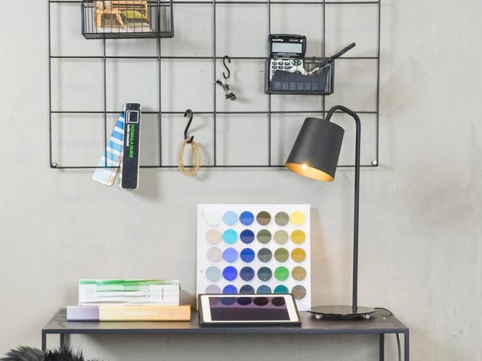 Et skrivebord med bøger, en åben tablet, en lampe og en farvepalet; bagved er der en metalgittervæg med kontorartikler og en lommeregner.
