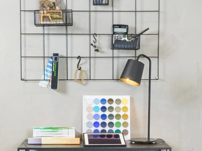 Et moderne skrivebord med en lampe, en farvepalet, bøger og en tablet under en trådgittervæg med forskellige kontorartikler.