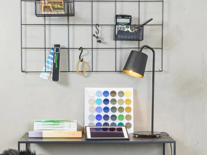 Et skrivebord med bøger, en åben tablet, en lampe og en farvepalet; bagved er der en metalgittervæg med kontorartikler og en lommeregner.