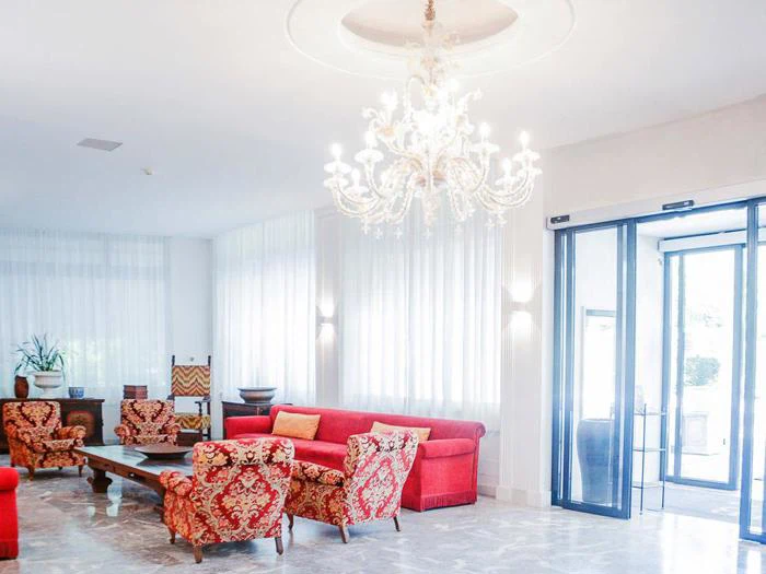 Lys hotellobby med røde og guldmønstrede sofaer, en stor lysekrone, marmorgulv og indgangsdøre af glas. Naturligt lys fylder rummet gennem rene, hvide gardiner.