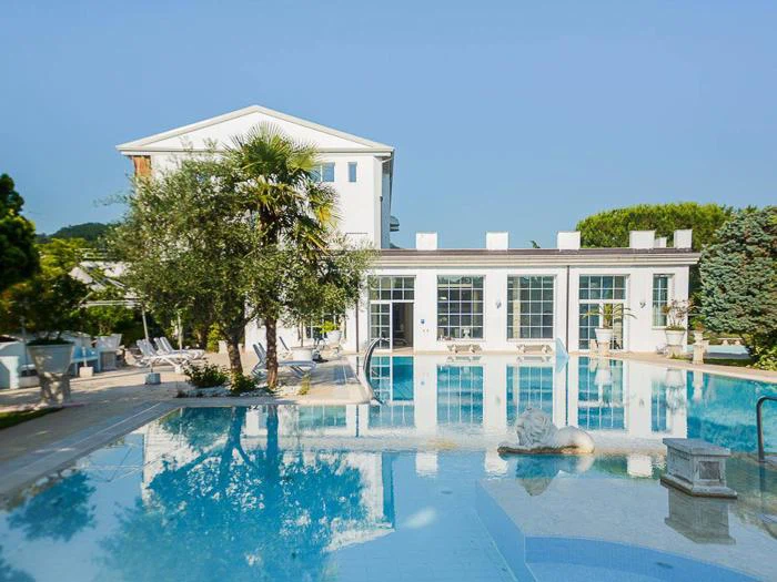 En klar udendørs swimmingpool med en løvestatue foran en moderne hvid bygning, omgivet af træer og liggestole på en solrig dag.