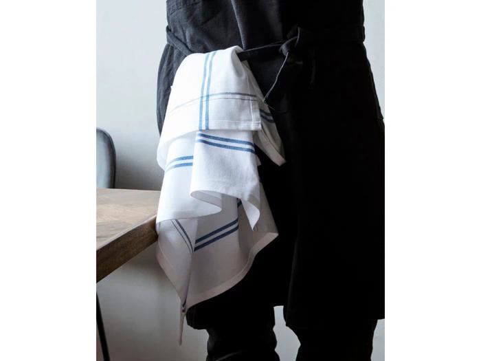 En person iført mørkt forklæde står ved siden af et træbord med et hvidt køkkenhåndklæde med blå striber stukket ind i forklædet.