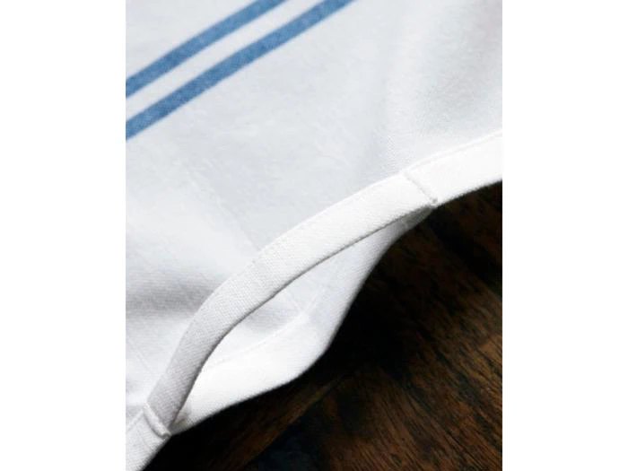 Nærbillede af hjørnet af et hvidt klæde med blå striber, der viser den syede kant, som ligger på en mørk træoverflade.