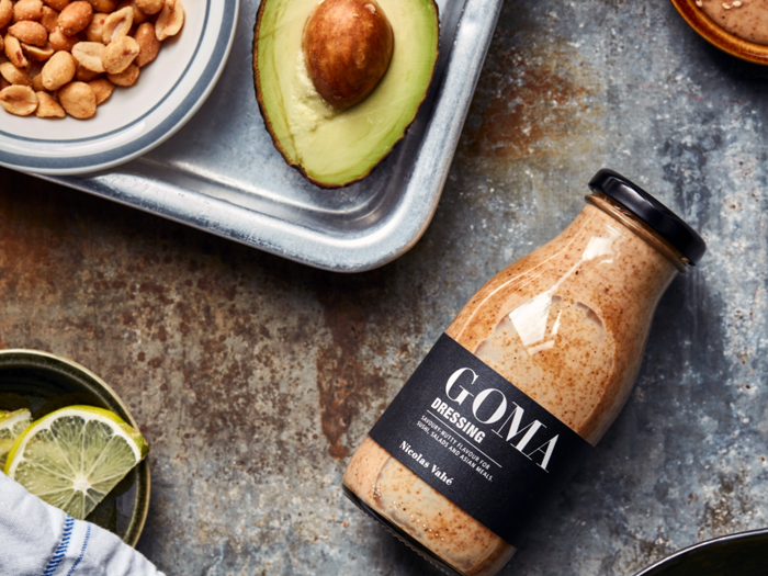 En flaske GOMA-dressing ligger på et bord ved siden af en halv avocado, en skål med peanuts og en tallerken med limeskiver.