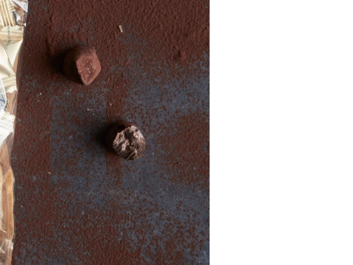 To chokoladetrøfler på en mørk, kakaostøvet overflade med en delvist synlig indpakning i venstre kant; højre side af billedet er blank hvid.